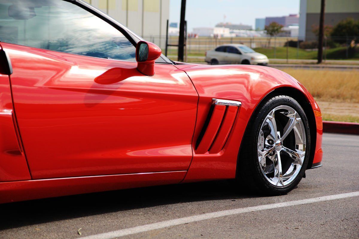Red Corvette sports car parked on asphalt. Chrome wheels, side air vents, and a side mirror are visible.