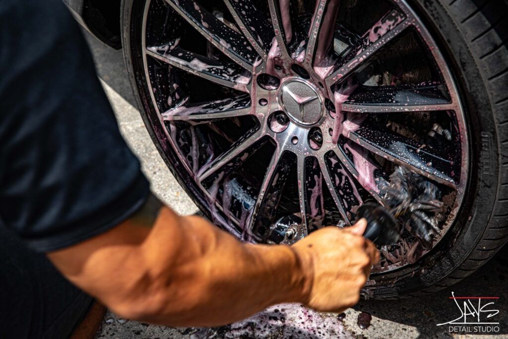 Person washes a black car wheel with pink soap, outdoors.