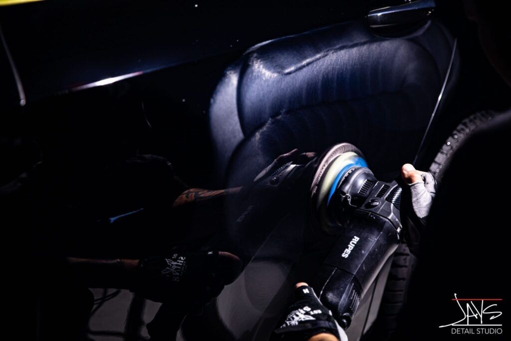 A person polishing a black car with a rotary buffer, creating a swirl pattern under a spotlight.