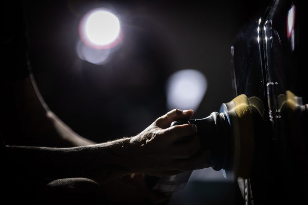 Hands polishing a car wheel in a dark setting, bright light in the background.