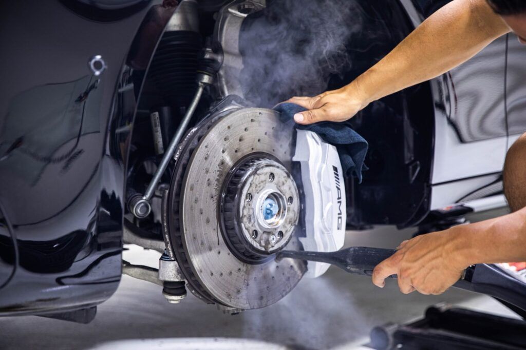 Person spraying steam cleaner on car brake rotor. Black car, silver rotor, blue caliper.