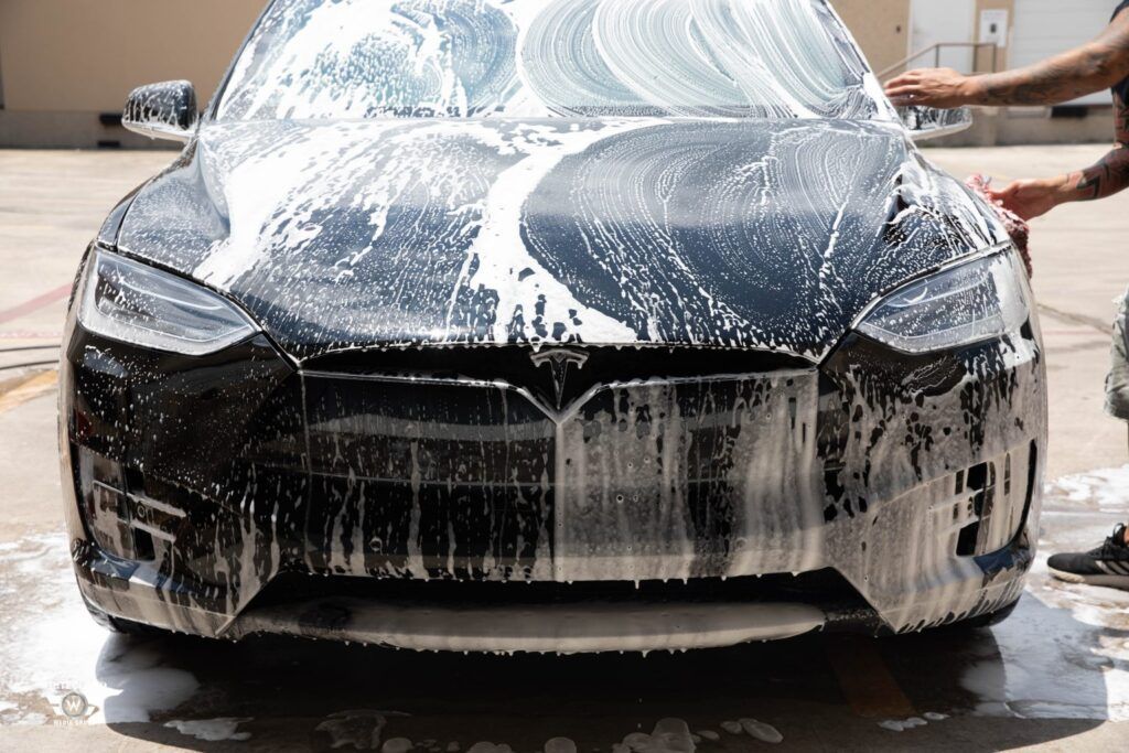 Black Tesla car being washed with foamy soap outdoors.