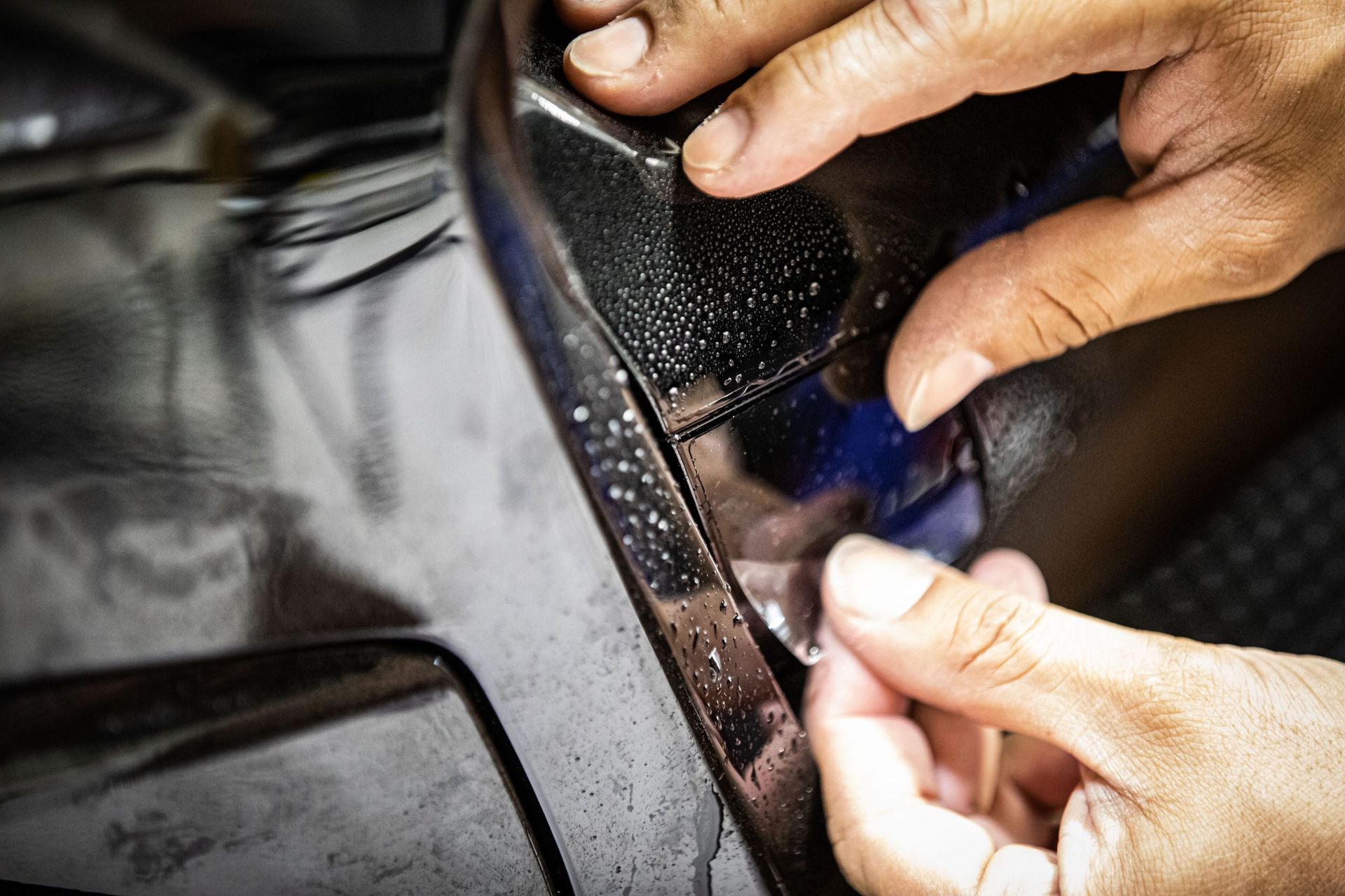 Hands applying protective film to a black car bumper.