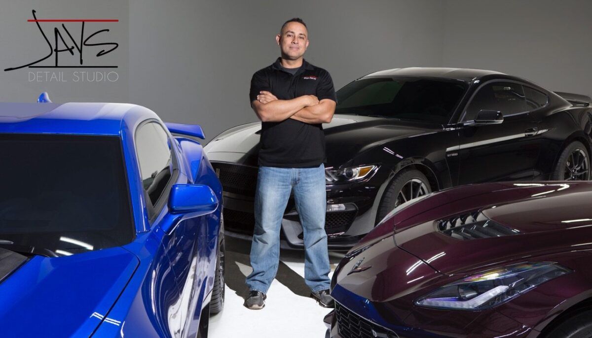 Man in black shirt and jeans stands with arms crossed, surrounded by three sports cars.