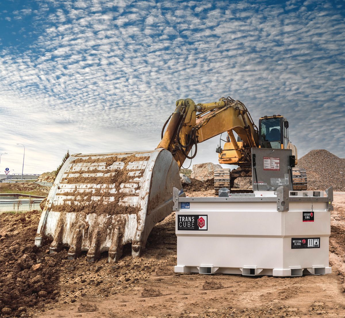Excavator working on a construction site next to a white industrial tank, under a cloudy sky.