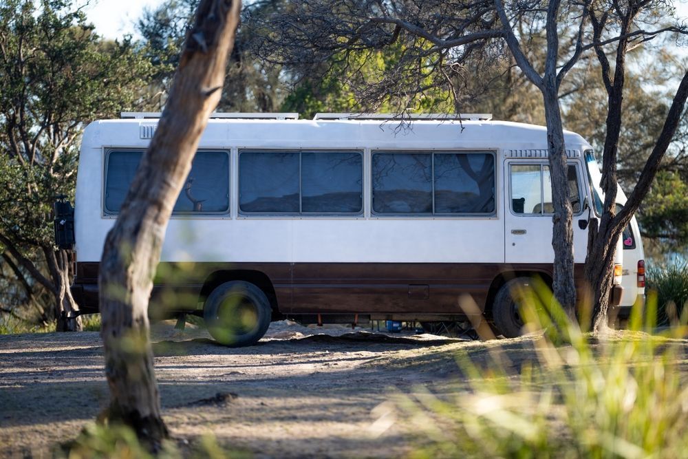A White Bus is Parked in a Dirt Lot Surrounded by Trees — Ozzie Camper Trailers in Aitkenvale, QLD