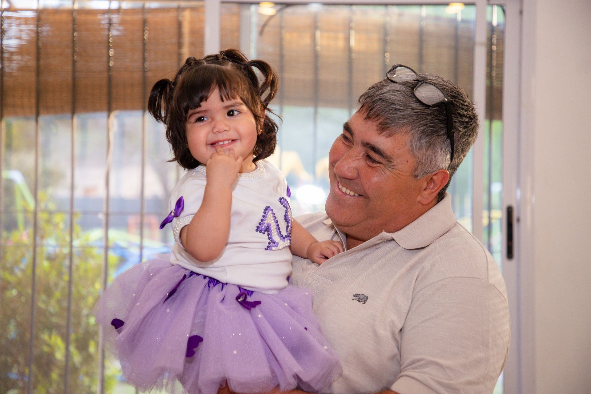 Un hombre sonriente sostiene a una joven con un tutú morado. Ambos están en casa, con aspecto feliz.