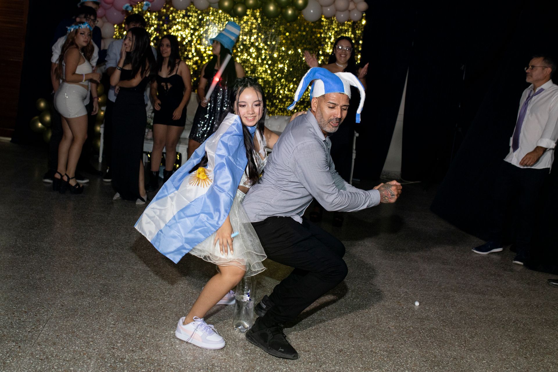 Una mujer y un hombre jóvenes bailan en una fiesta, luciendo sombreros novedosos y una bandera argentina.