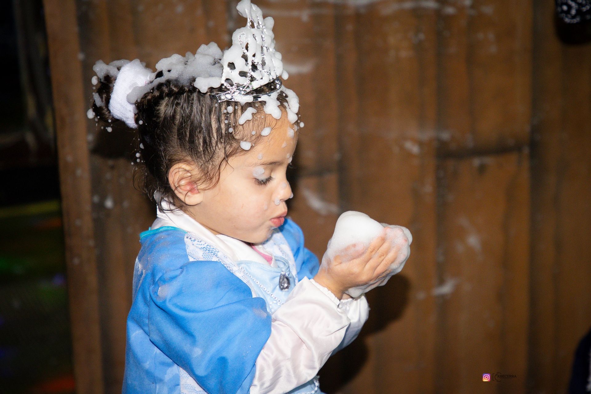 Niña con corona de espuma, soplando espuma con las manos. Lleva vestido azul y blanco sobre fondo de madera.
