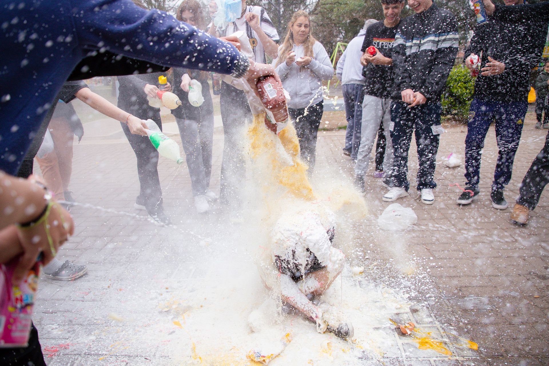 Personas arrojando harina y otras sustancias sobre alguien que yace en el suelo, al aire libre.