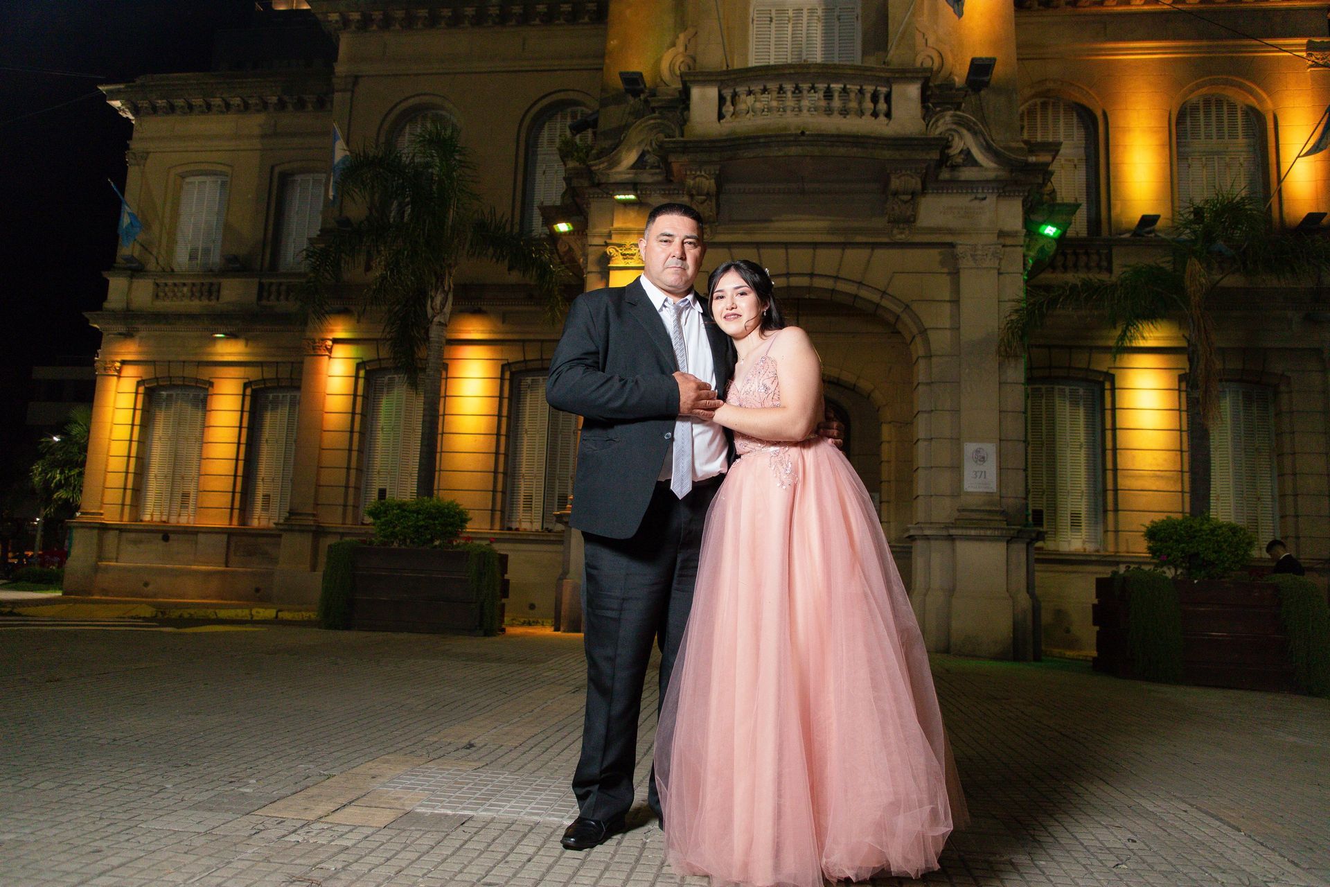 Pareja en traje formal posando frente a un edificio iluminado por la noche.