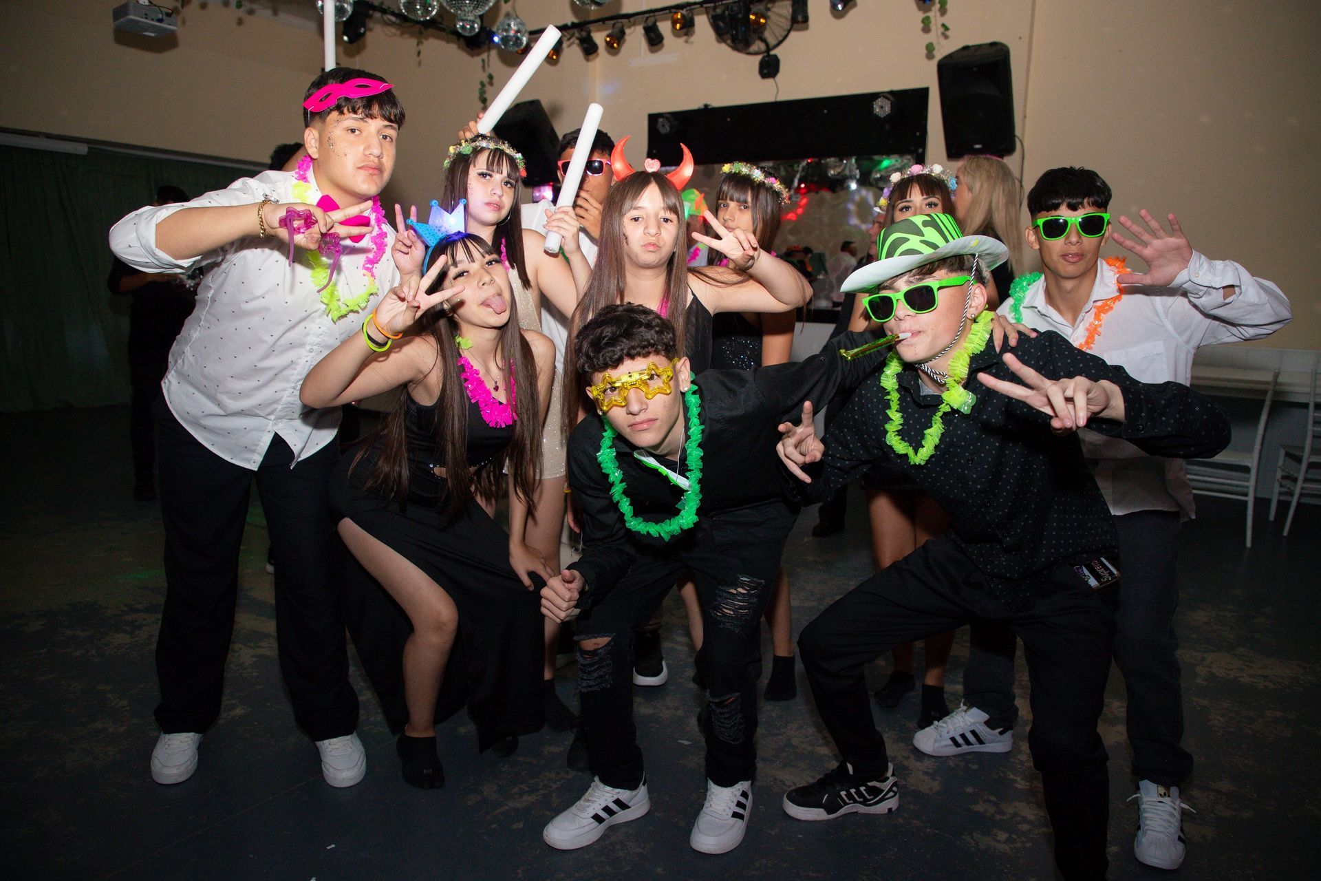Grupo de adolescentes posando en una fiesta, algunos con accesorios de neón y haciendo señales de paz.