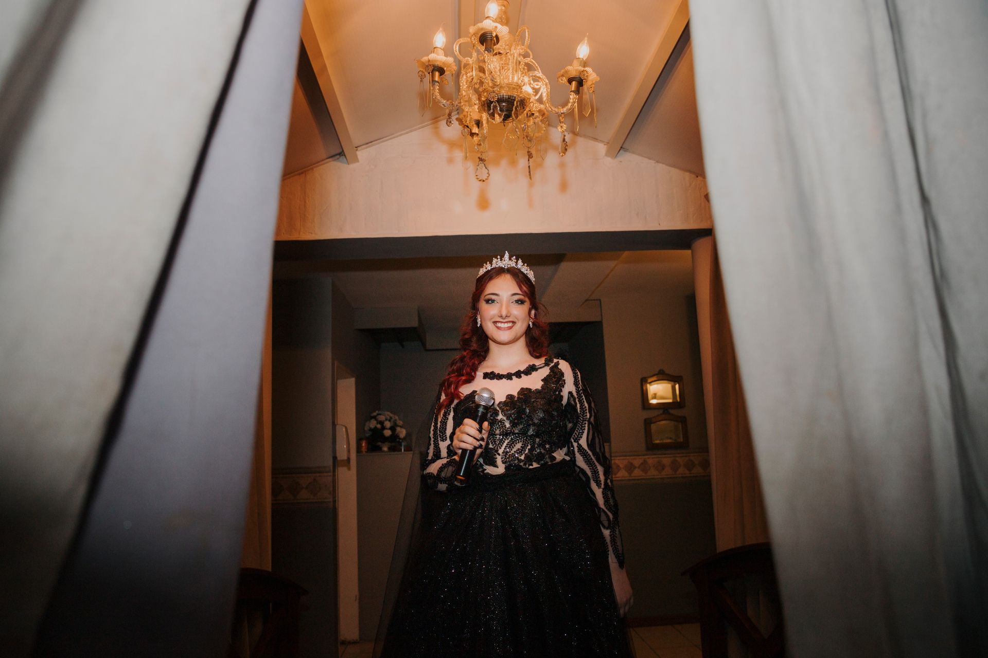 Una mujer con un vestido negro y una tiara, sosteniendo un vaso, está parada en la puerta debajo de una lámpara de araña.