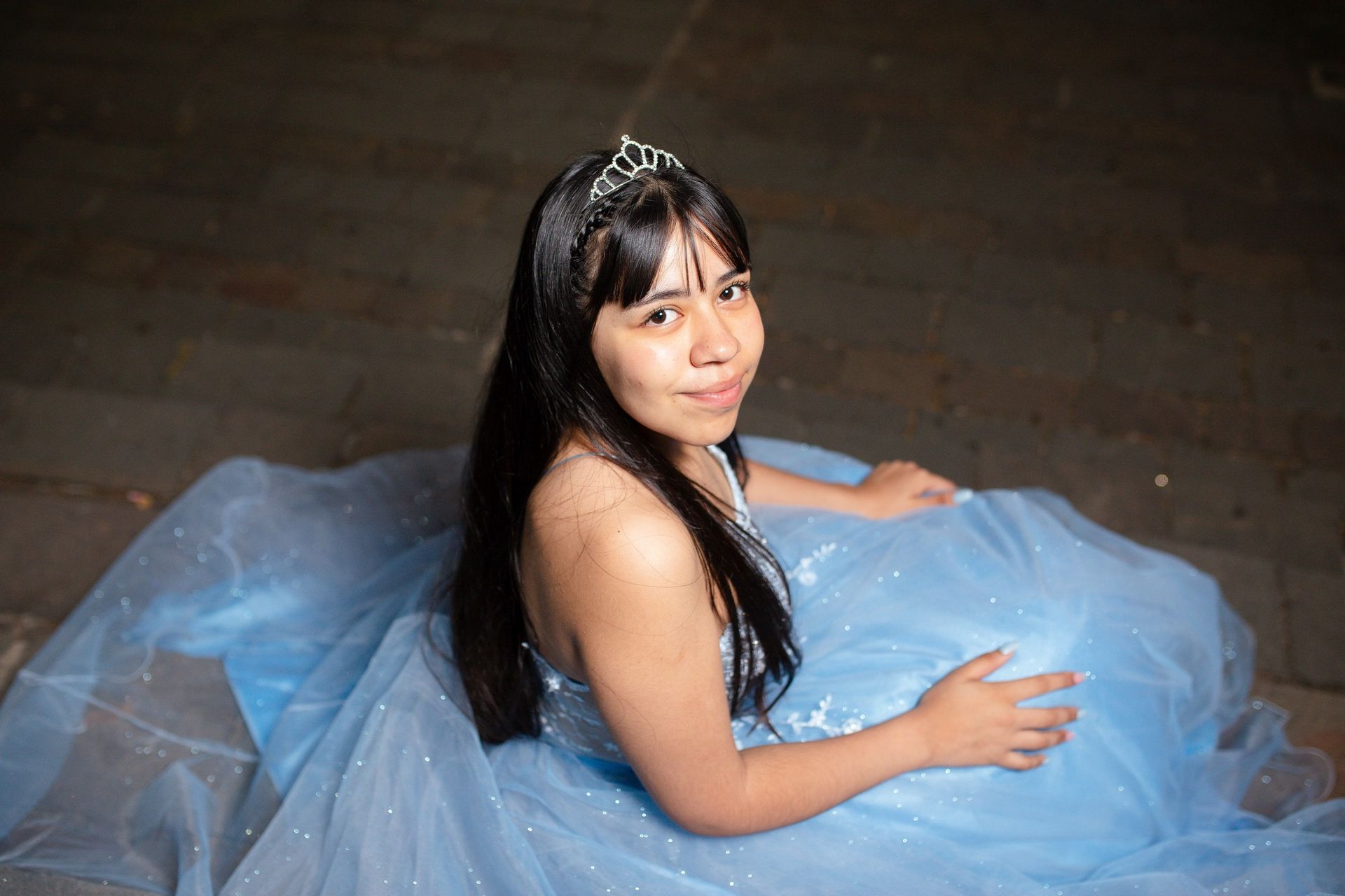 Mujer con vestido azul claro y tiara, mirando hacia arriba con una leve sonrisa. Escenario al aire libre.