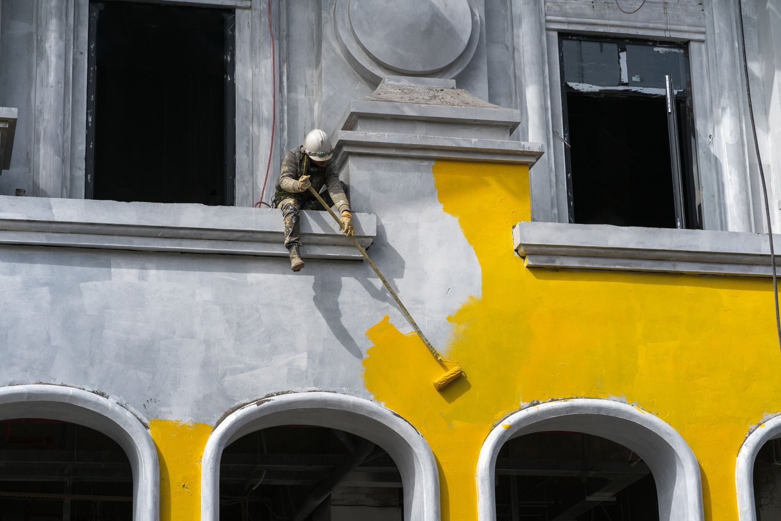 Painter in yellow overalls using a roller to paint building facade bright yellow.
