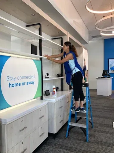 Woman on a stepladder arranging shelves in a store. White shelves, blue walls, a sign reads, 