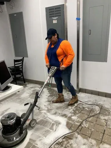Person in orange shirt and apron using a floor cleaner in a room with tiled flooring.
