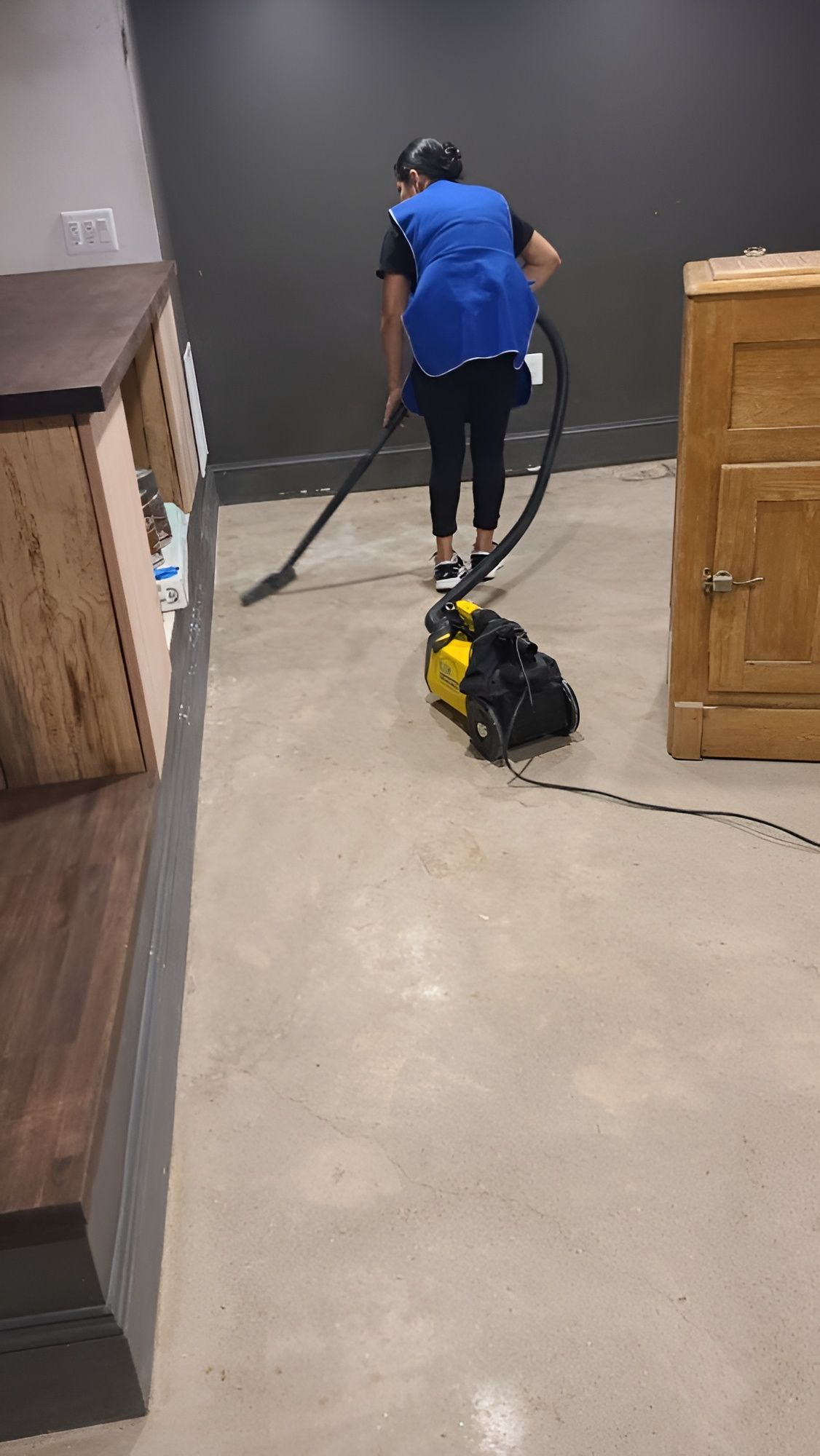 Person vacuuming a floor near wooden cabinets in a room with gray walls.