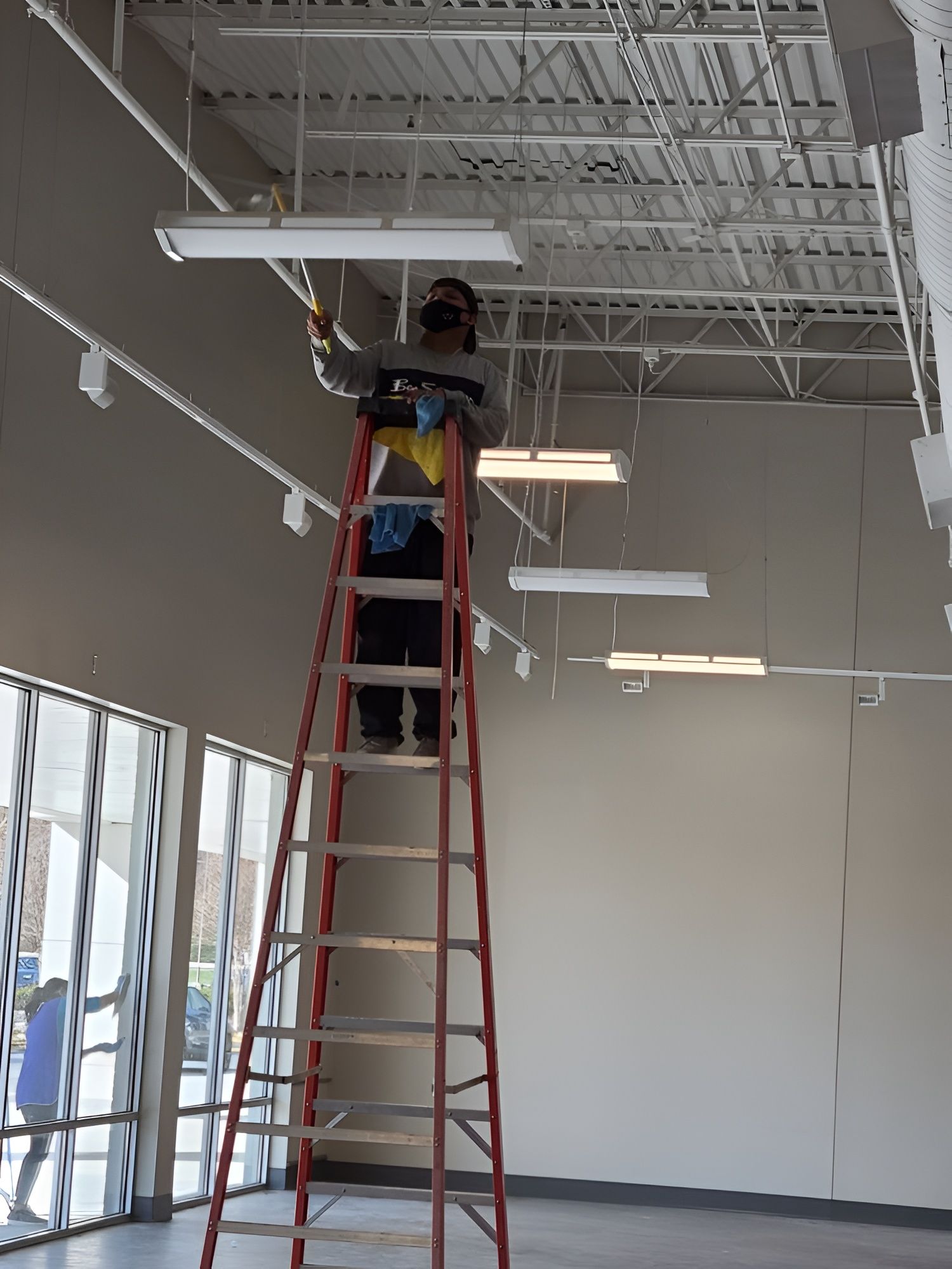 Person on a ladder cleaning a light fixture in a large room with high ceilings.