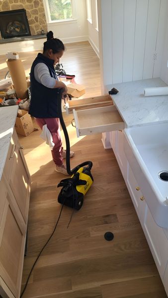 Person vacuuming a cabinet drawer in a kitchen with light-colored wooden floors and white cabinets.
