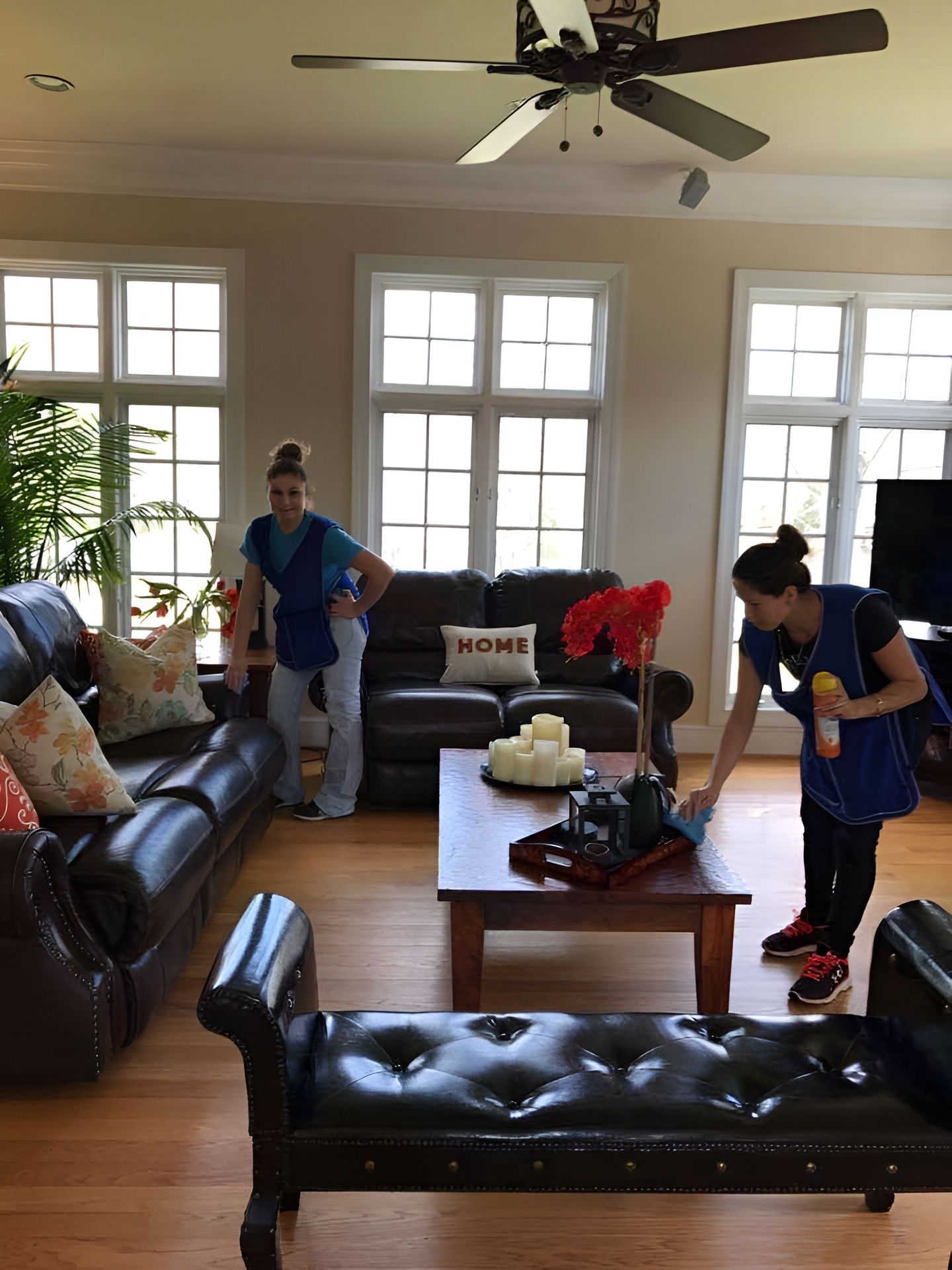 Two people cleaning a living room with dark leather furniture and large windows.