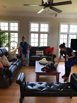 Two people cleaning a living room with dark leather furniture and large windows.