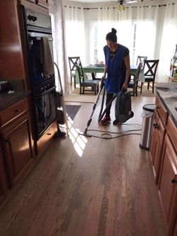 Person vacuuming hardwood floor in a kitchen, wearing a blue apron, with a dining area in the background.