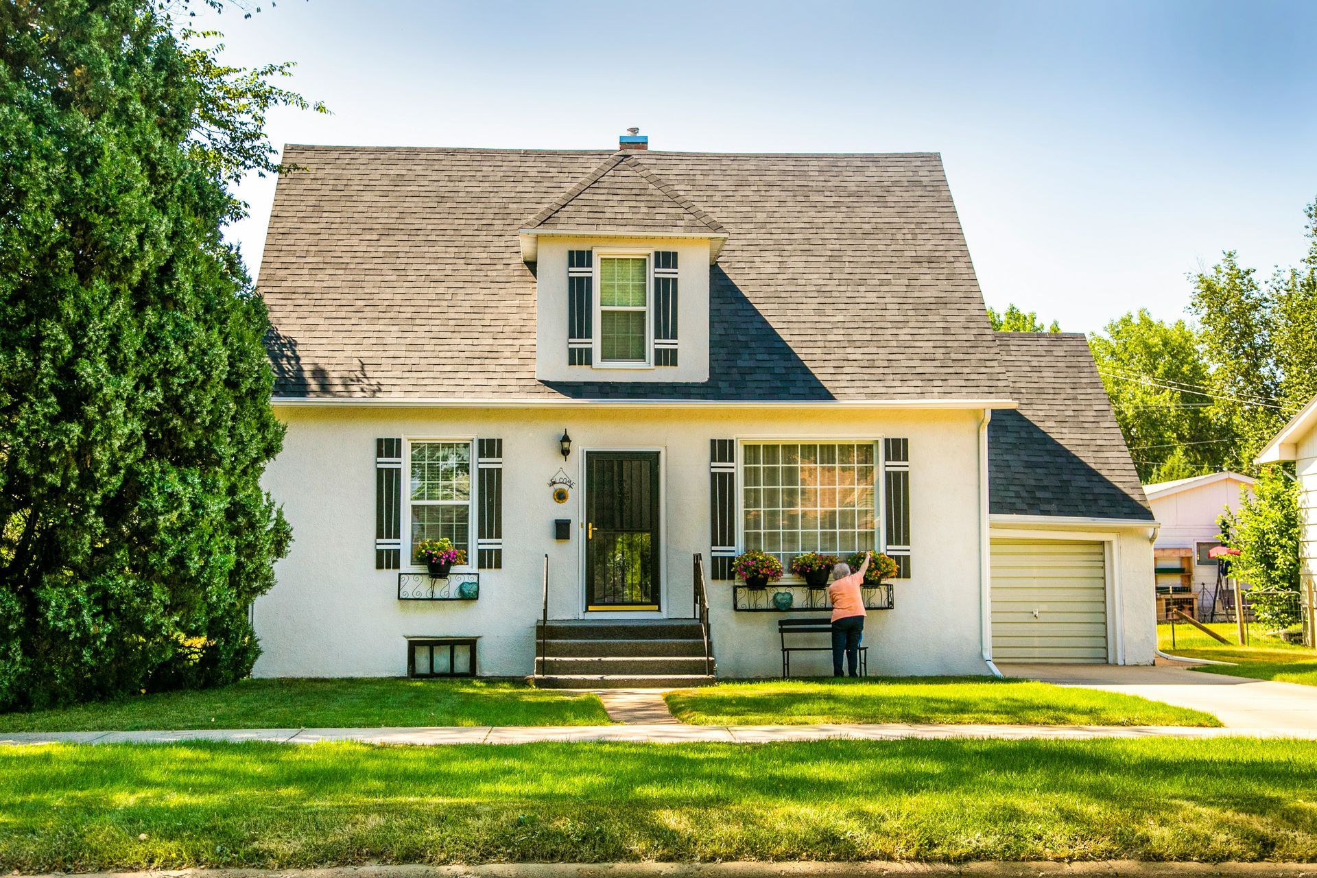 A white house with a gray roof and dormer window, surrounded by green lawns and trees, with a person gardening out front.