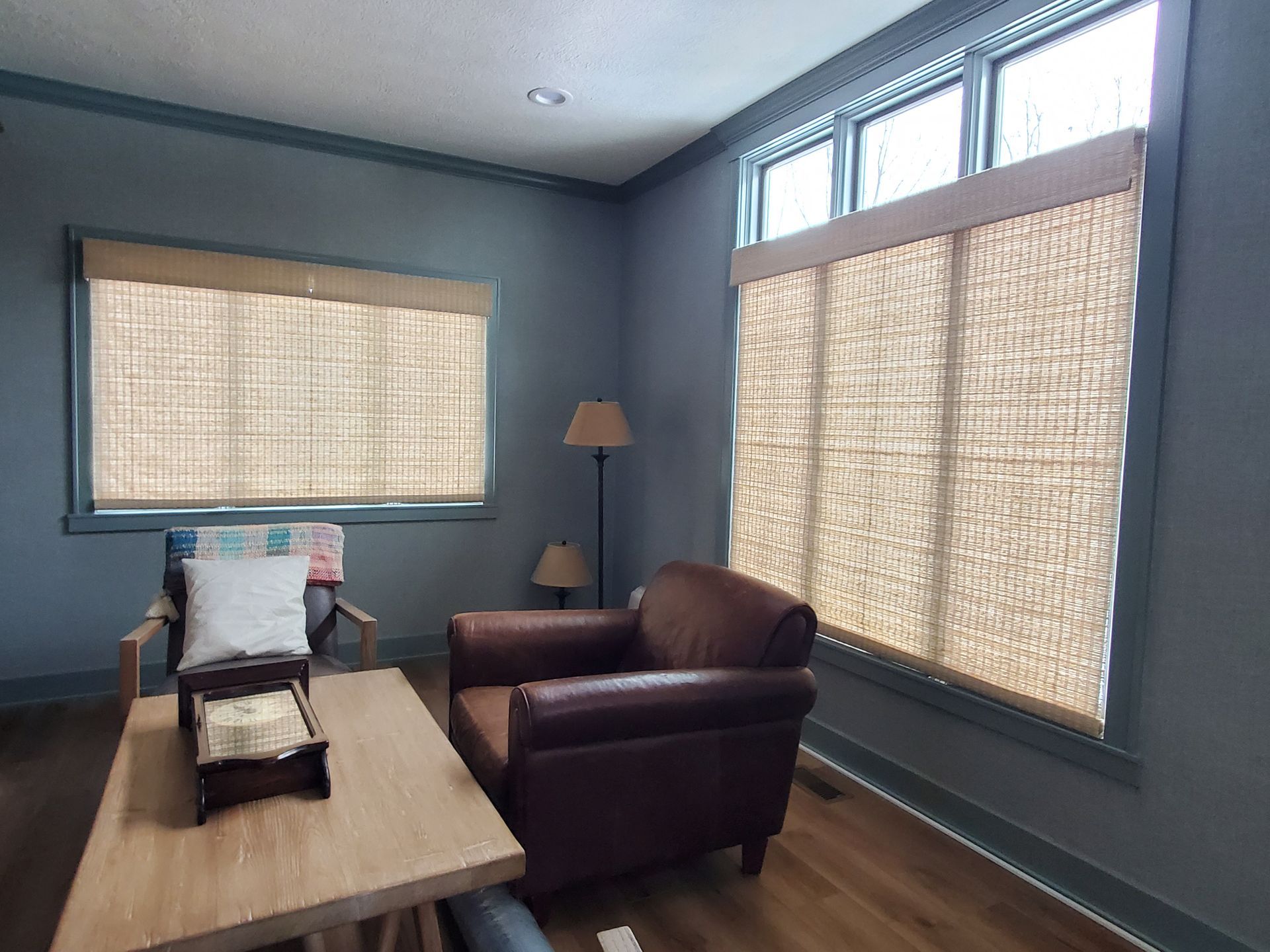 Two windows, both trimmed in dark blue and set into dark blue walls, are fitted with light-colored woven wood shades. The shades are lowered to cover the majority of both windows. Through the uncovered top portion of the right window, a bright, overcast sky is visible. In the foreground, a wooden coffee table sits between a light-colored armchair with a patterned throw and a dark brown leather armchair. A floor lamp with two beige shades is visible in the corner behind the leather armchair.