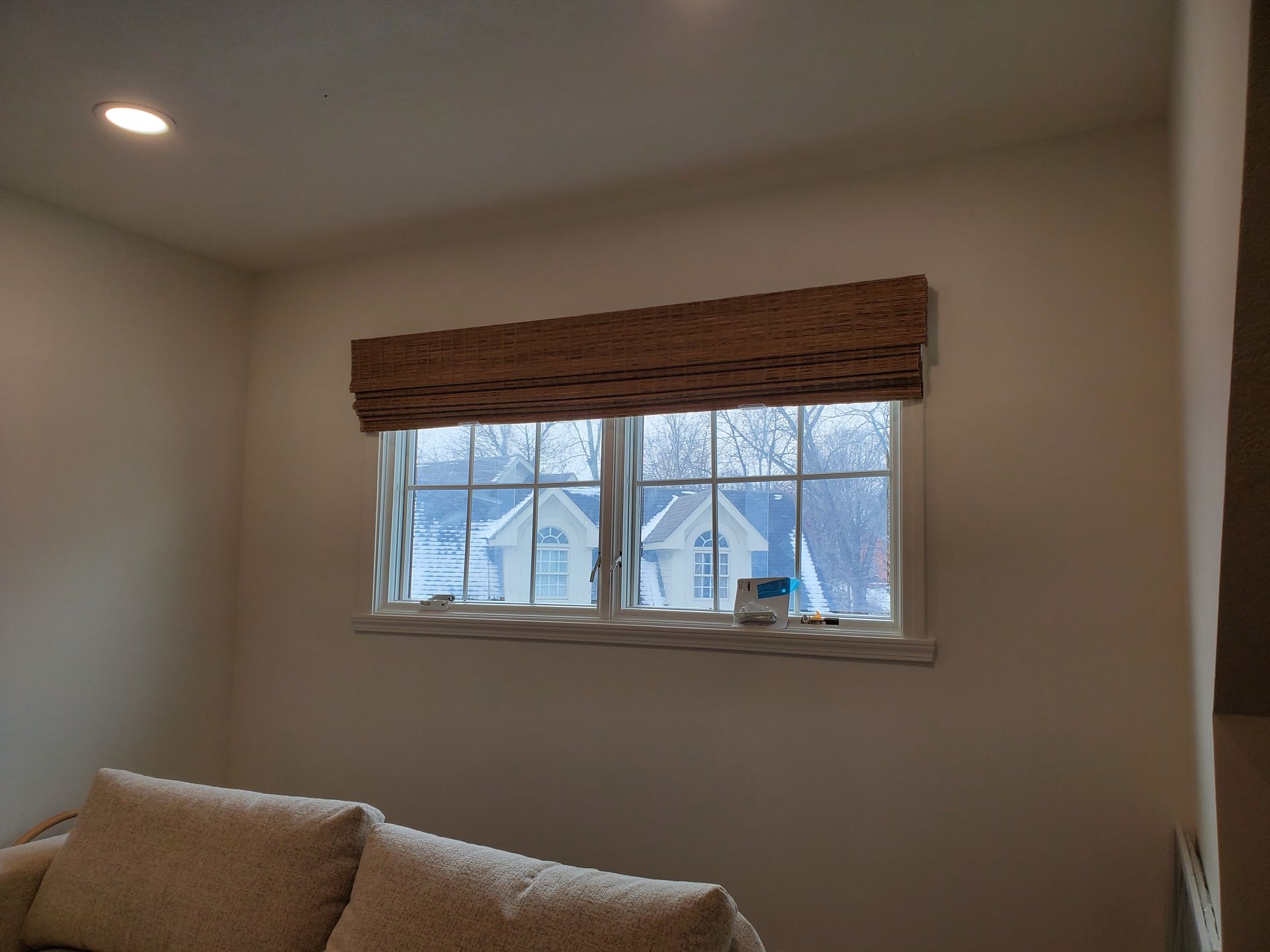 A horizontally oriented window, framed in white, is partially covered by a flat-fold woven wood shade in a light brown color. The shade is raised to reveal the lower half of the window, through which two white-gabled houses with dark roofs and snow-covered ground are visible. The walls of the room are a light, neutral color, and a recessed light is visible in the ceiling. A portion of a light-colored sofa or sectional is in the foreground.