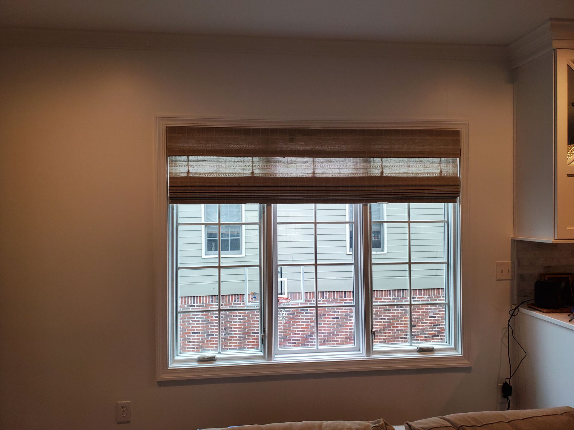 A triple window unit is partially covered by a woven wood shade in a light brown tone, lowered to conceal the top portion of the window. The uncovered lower sections of the windows reveal the exterior of a neighboring building with light-colored siding and red brick. The room's walls are a light neutral color. A small section of a light-colored sofa is visible in the bottom right corner, and a white cabinet with a glass front is partially visible on the far right.