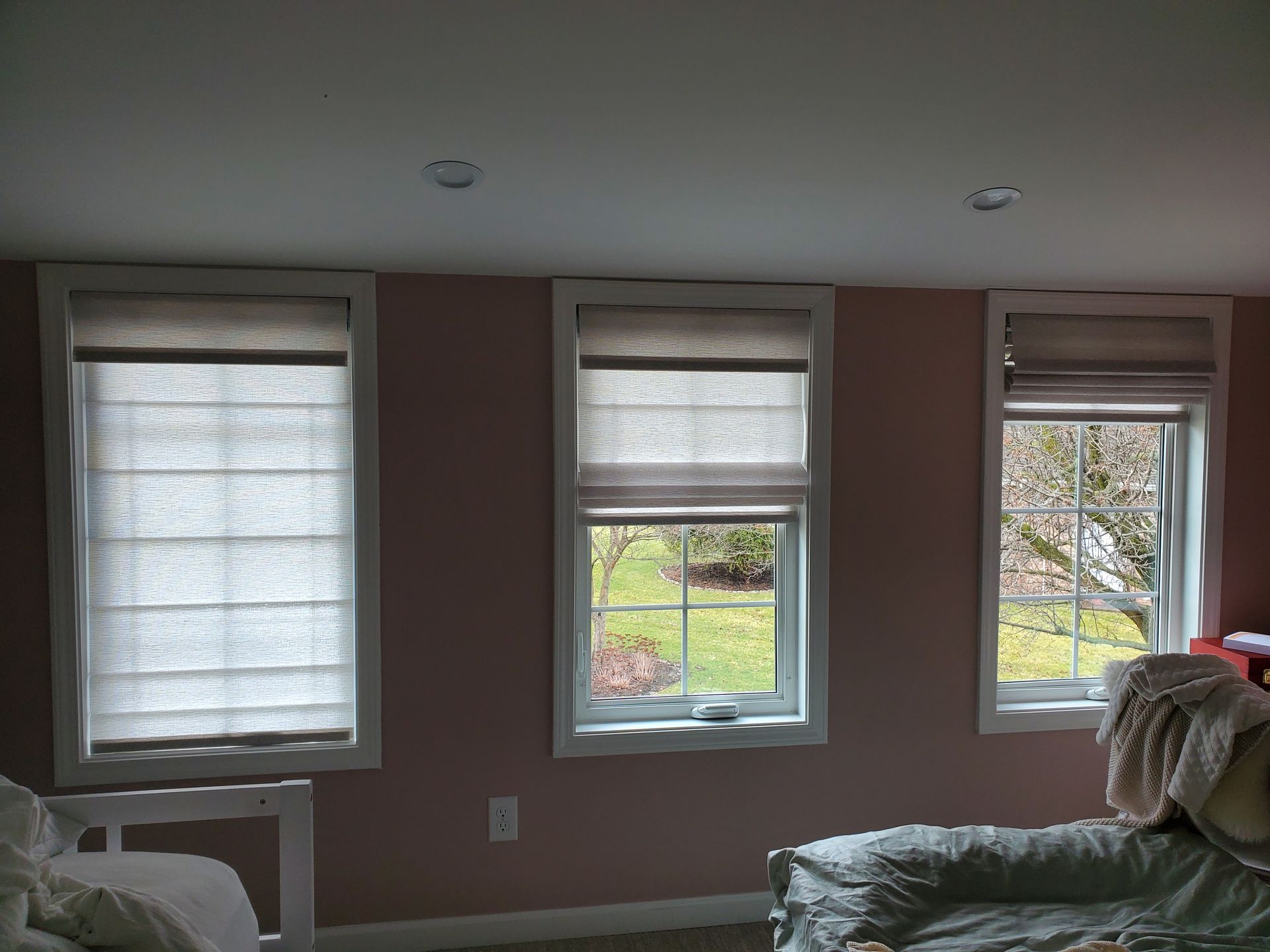 Three windows with white trim are set against a light pink wall. Each window features two-tone roman shades: the top portion is a darker tan, while the lower, larger section is a lighter, semi-sheer white, allowing some light to filter through. The shades on the left and middle windows are pulled down, while the roman shade on the right window is pulled up slightly, revealing a partial view of a green lawn and trees outside. A corner of a white bed frame is visible on the far left, and a bed with a rumpled light green duvet is on the far right. Two recessed lights are visible in the ceiling.