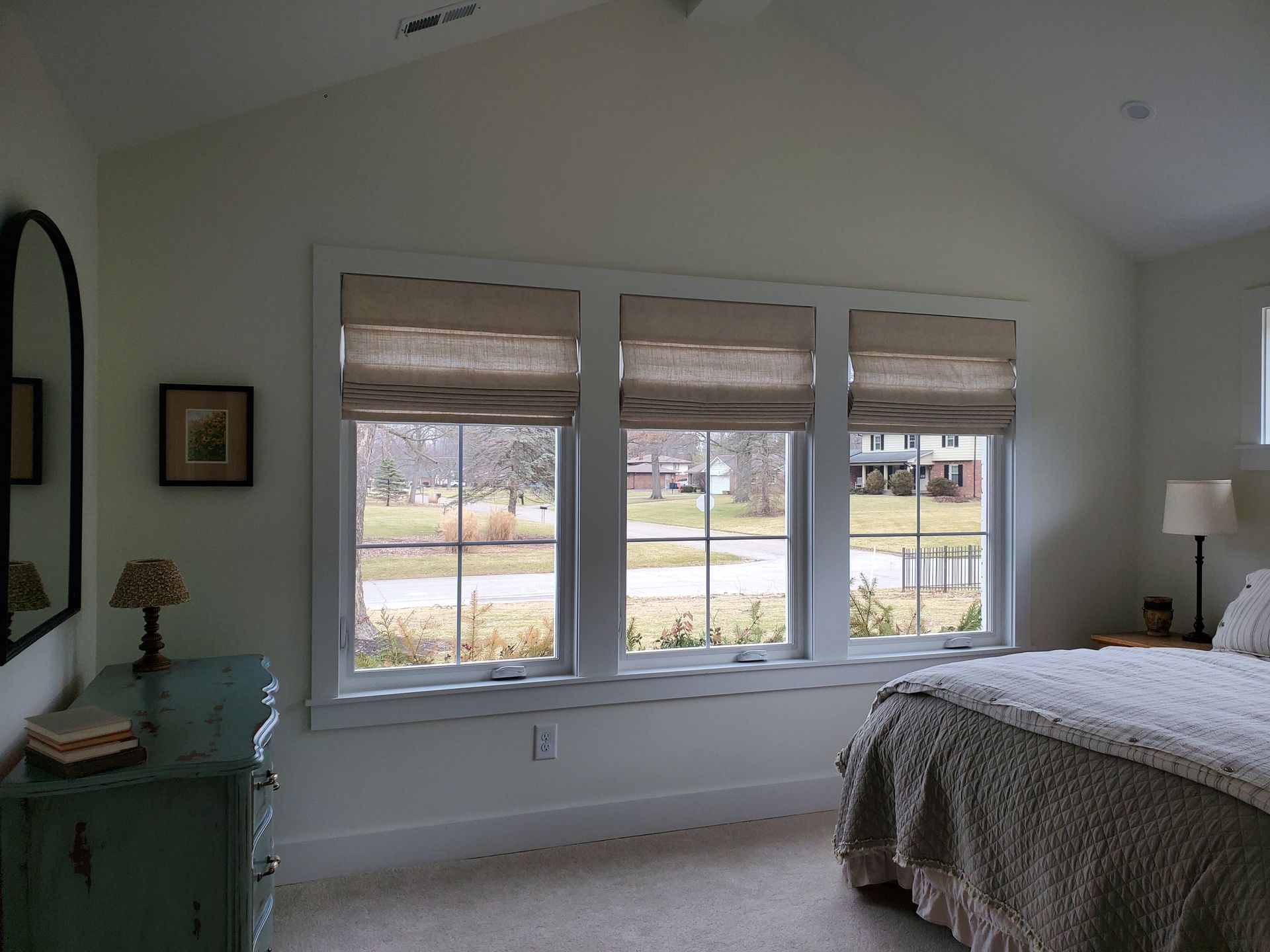 Three double-hung windows with white trim are installed in a light-colored wall, possibly a bedroom. Each window is adorned with tan roman shades pulled part-way down, leaving the bottom half of the windows exposed. Through the windows, a suburban street scene is visible with bare trees, grass, and houses in the distance, indicating a view of a neighborhood. To the left, a dark-framed mirror hangs above a distressed light blue dresser, on which sits a small lamp and a stack of books. On the right, a bed with a neutral-colored comforter and a lamp on a nightstand is partially visible.