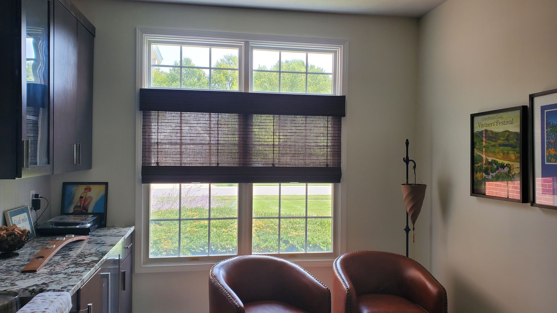 A large window with white framing is partially covered by dark brown woven wood shades, which are lowered to cover the middle section of the window. The top and bottom sections of the window are uncovered, revealing a sunny outdoor scene with green grass, trees, and what appears to be a residential area in the distance. To the left of the window, dark brown cabinets with glass inserts are visible above a countertop. In the foreground, two brown leather armchairs are positioned facing the window. On the right wall, two framed art pieces are hanging.