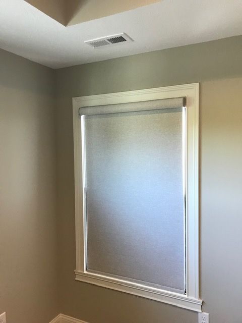 A single window is fitted with a light gray roller-style room darkening shade, fully extended to cover the entire window. The window has white trim and is set into a light beige wall. A white air vent is visible on the ceiling above the window.