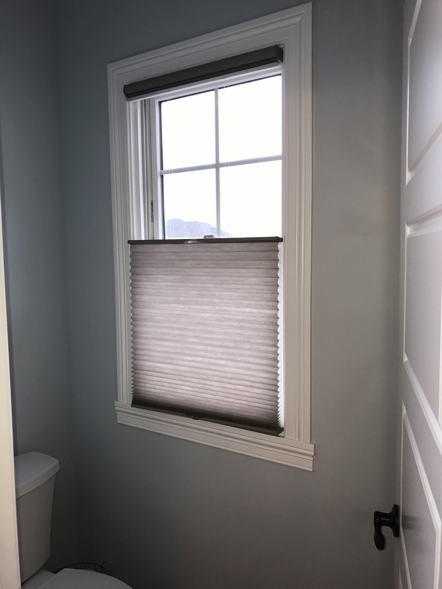 A white-framed window in a room with light gray walls features light-filtering cellular shades in a top-down, bottom-up configuration. The lower portion of the window is covered by the cellular shades, while the top portion reveals a bright, overcast sky outside. A white toilet tank is visible in the lower left, and a white paneled door with a black handle is partially open on the right.