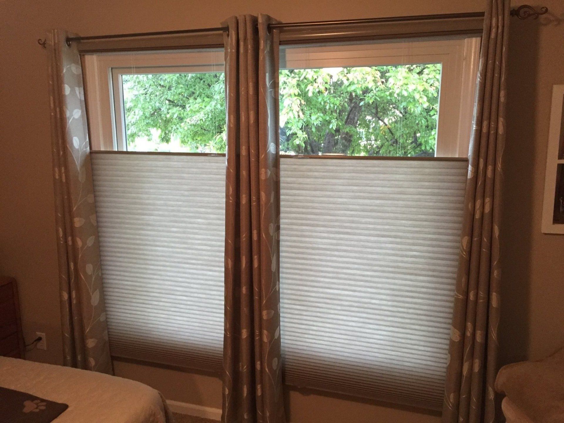 Two large windows are adorned with light-colored cellular shades, extended from the bottom up to cover the lower two-thirds of each window. The top portion of the windows reveals the lush green leaves of a tree outside. On either side of each window, brown and beige patterned curtains are pulled back and held by tie-backs. A bed or furniture is partially visible in the bottom left corner.