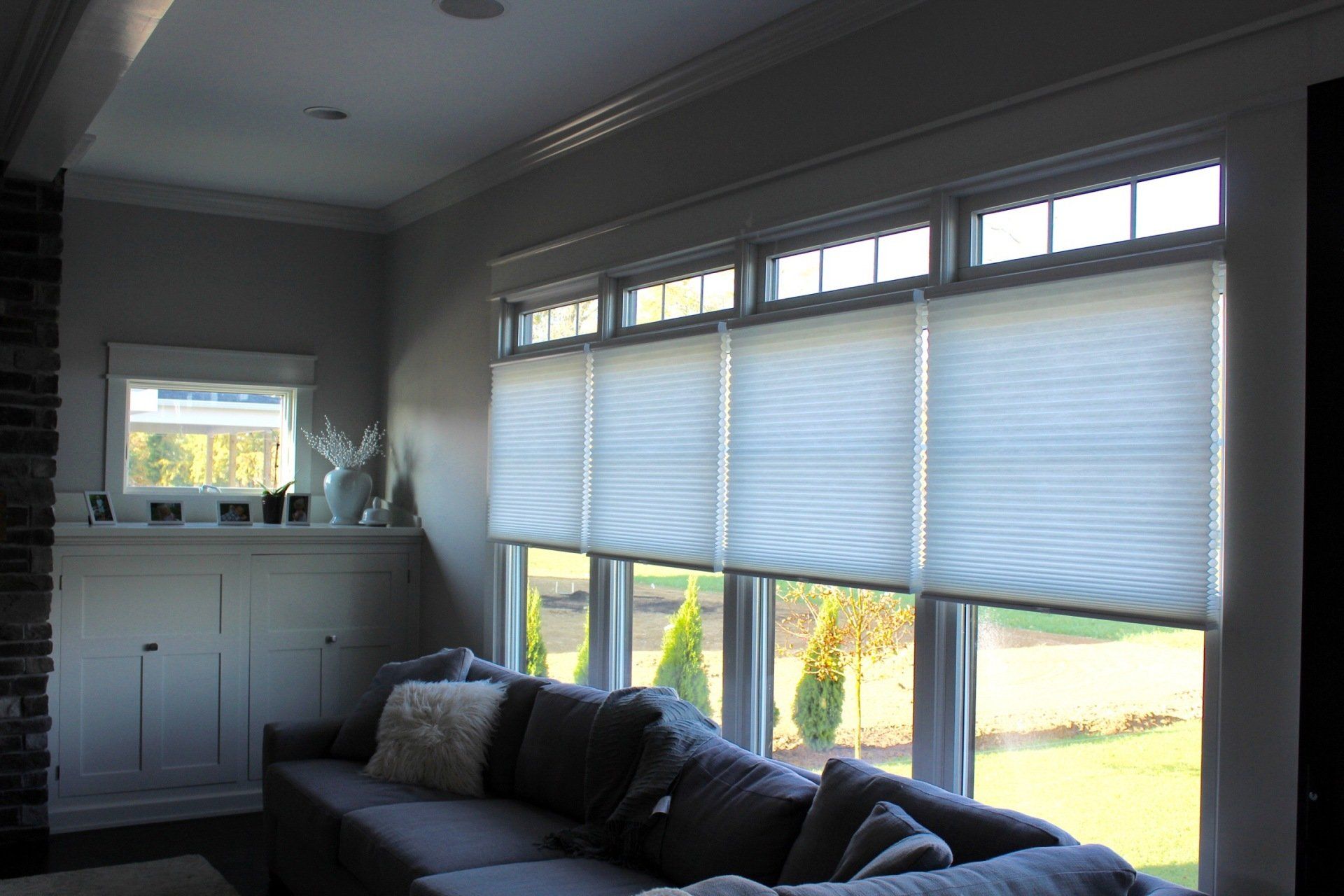 A wide bank of windows in a living room is equipped with light-filtering cellular shades. The shades are pulled up from the bottom, leaving the top transom windows uncovered and allowing natural light to enter. A dark gray sectional sofa with a white throw pillow is in the foreground. To the left, a smaller window is visible above a white built-in cabinet, and a brick column stands in the far left. Outside, green grass and small evergreen trees are visible.