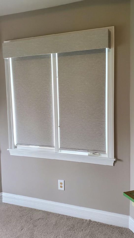 Two light gray room darkening shades are installed side-by-side within a white-trimmed window frame. 