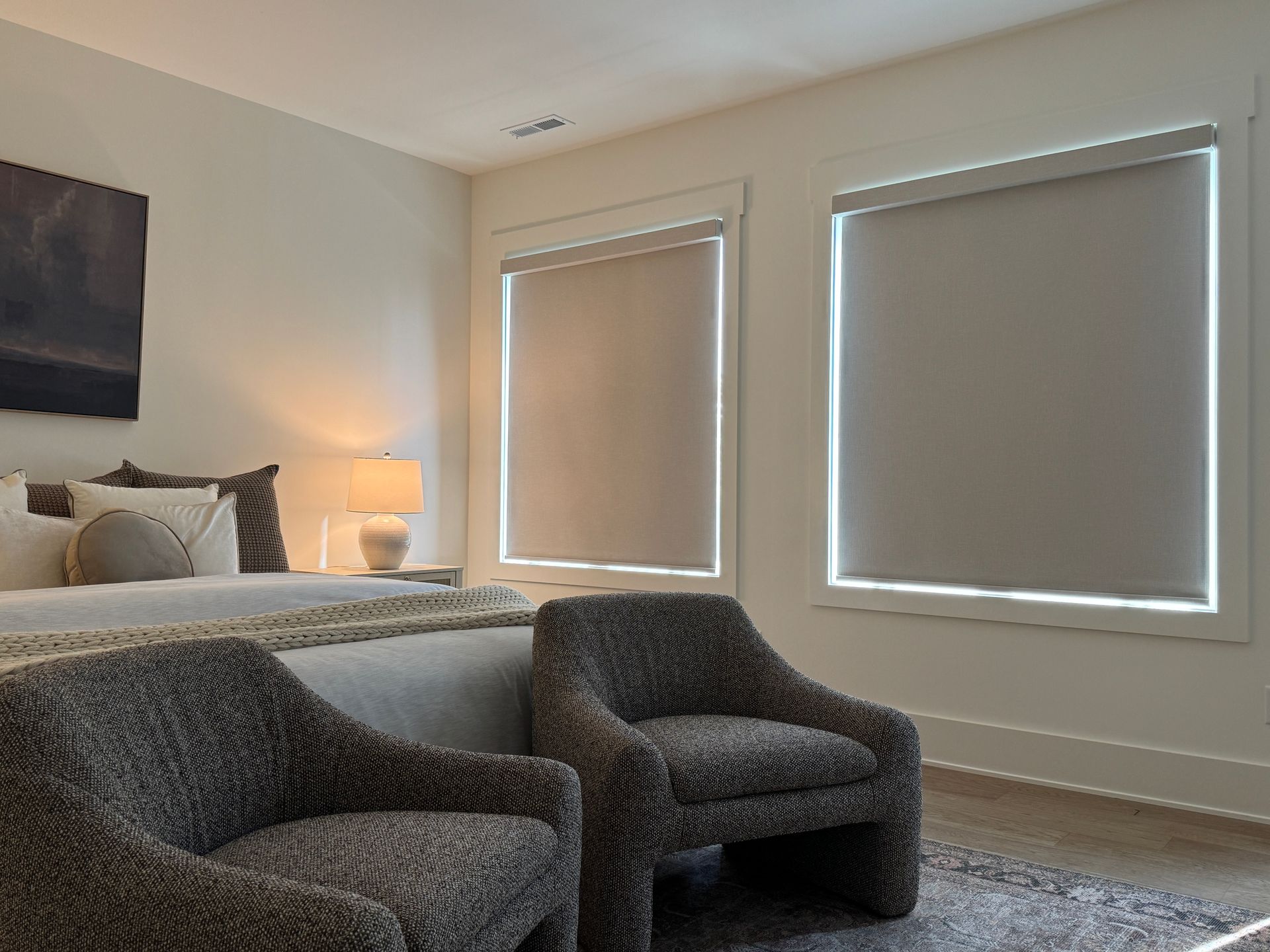 Two large windows in a bedroom are fitted with light gray room darkening shades, pulled down to cover the entire window and block light. The walls are a light neutral color. In the foreground, two textured gray armchairs sit on a patterned rug. A bed with a light-colored duvet and pillows is visible to the left, with a nightstand and a lit lamp beside it. A dark framed abstract art piece hangs above the bed.