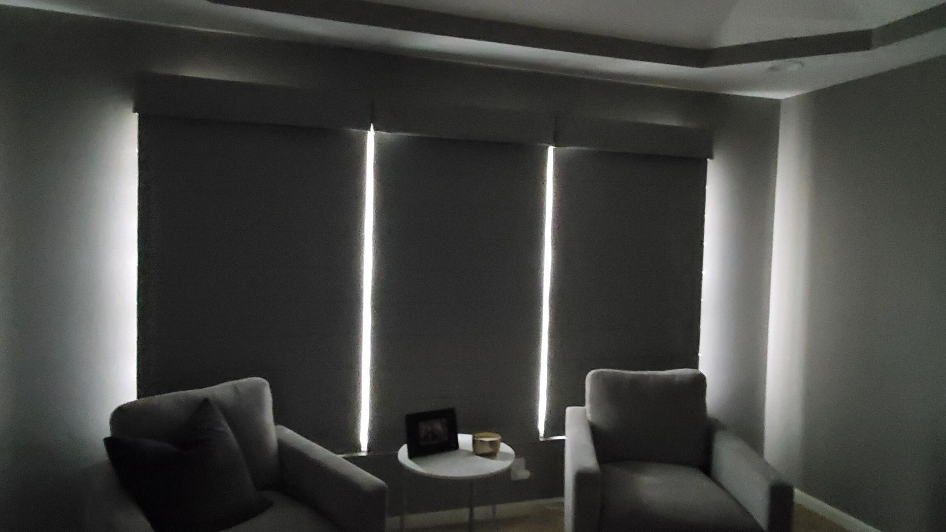 Two windows are fitted with dark gray room darkening shades, which are fully extended, allowing only thin vertical lines of light to escape from the sides. Two light-colored upholstered armchairs face the viewer, with a small round white side table between them holding a framed picture and a decorative object. The room's walls are a dark gray, and the ceiling has a tray design with recessed lighting.