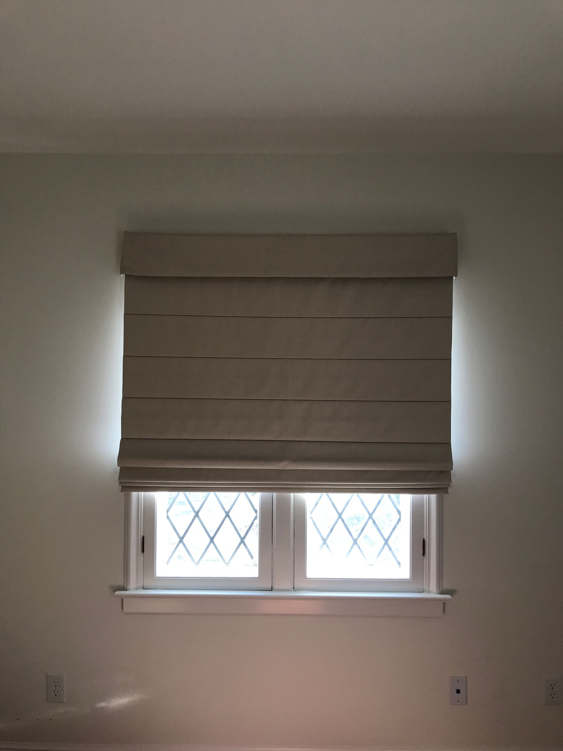A beige Roman shade, functioning as a room darkening shade, covers the upper portion of a window, allowing light to emanate from behind its edges. The lower section of the window features diamond-patterned grilles, suggesting multiple panes of glass. The window is set into a plain light-colored wall.