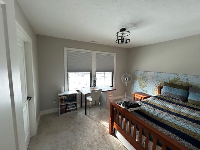 A bedroom features a light-colored carpet and gray walls. Two windows on the far wall are fitted with light gray room darkening shades, which are pulled down to cover the windows. A white desk with a chair is positioned in front of the windows, flanked by a small white bookshelf. To the right, a wooden bed frame with a striped comforter and a headboard featuring a world map print is visible. A white standing fan is next to the bed, and a black, caged ceiling light fixture hangs from the ceiling. A white door with a black handle is open on the left.