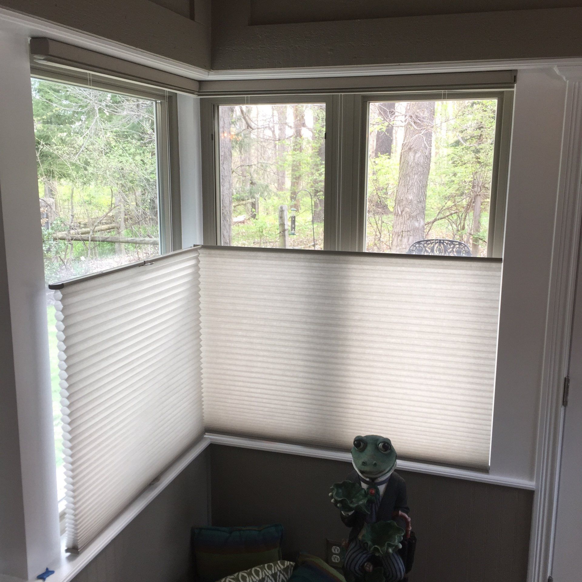 A set of windows forming a corner in a room is fitted with light-colored, pleated motorized shades in a top-down, bottom-up style. The shades cover the lower half of the windows, allowing views of green trees and bare branches through the upper sections. A decorative frog statue in a suit is visible in the foreground, and a patterned pillow is partially visible on what appears to be a bench or seating area below the window.