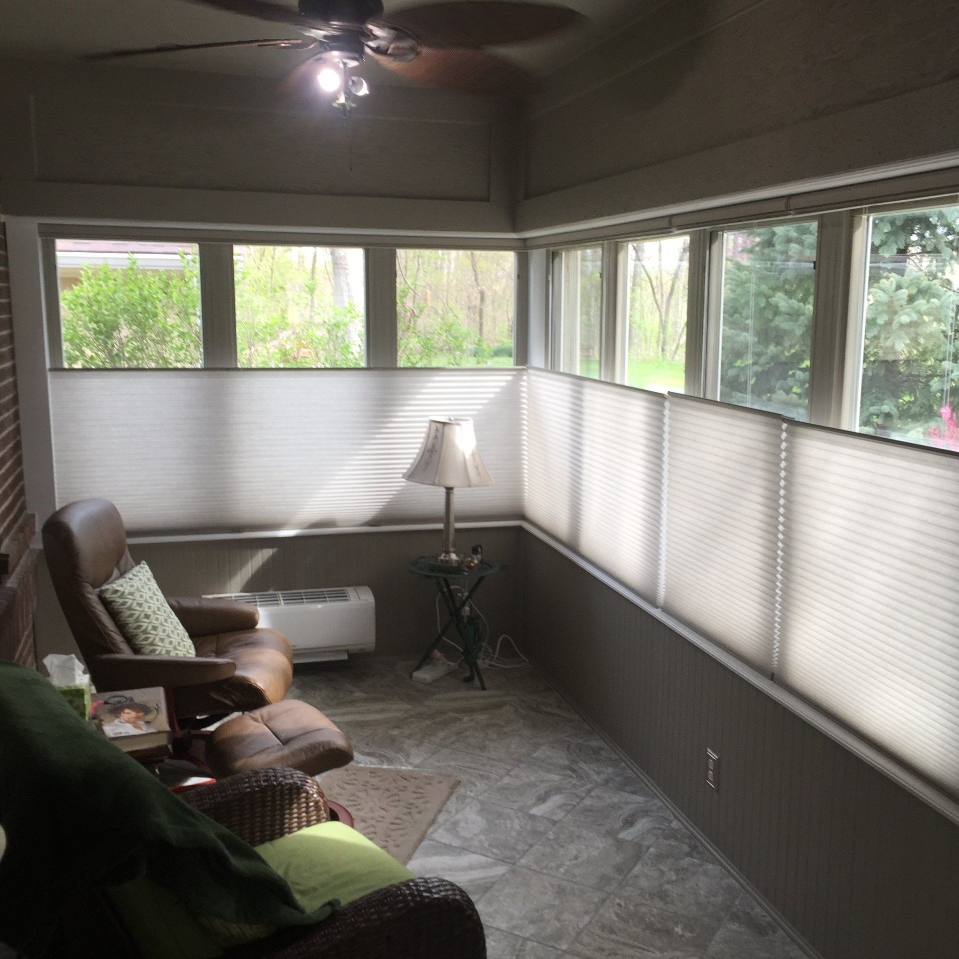 A sunroom or enclosed porch features numerous windows, all fitted with light-colored, pleated motorized shades. The shades are in a top-down, bottom-up configuration, covering the lower portion of the windows while leaving the top sections open to reveal green trees and foliage outside. The room has gray walls and a tiled floor. A brown leather reclining chair and ottoman are visible on the left, next to a white heater unit. A lamp with a white shade sits on a small table in the corner. Another chair with a green throw is partially visible in the foreground. A ceiling fan with lights is overhead.