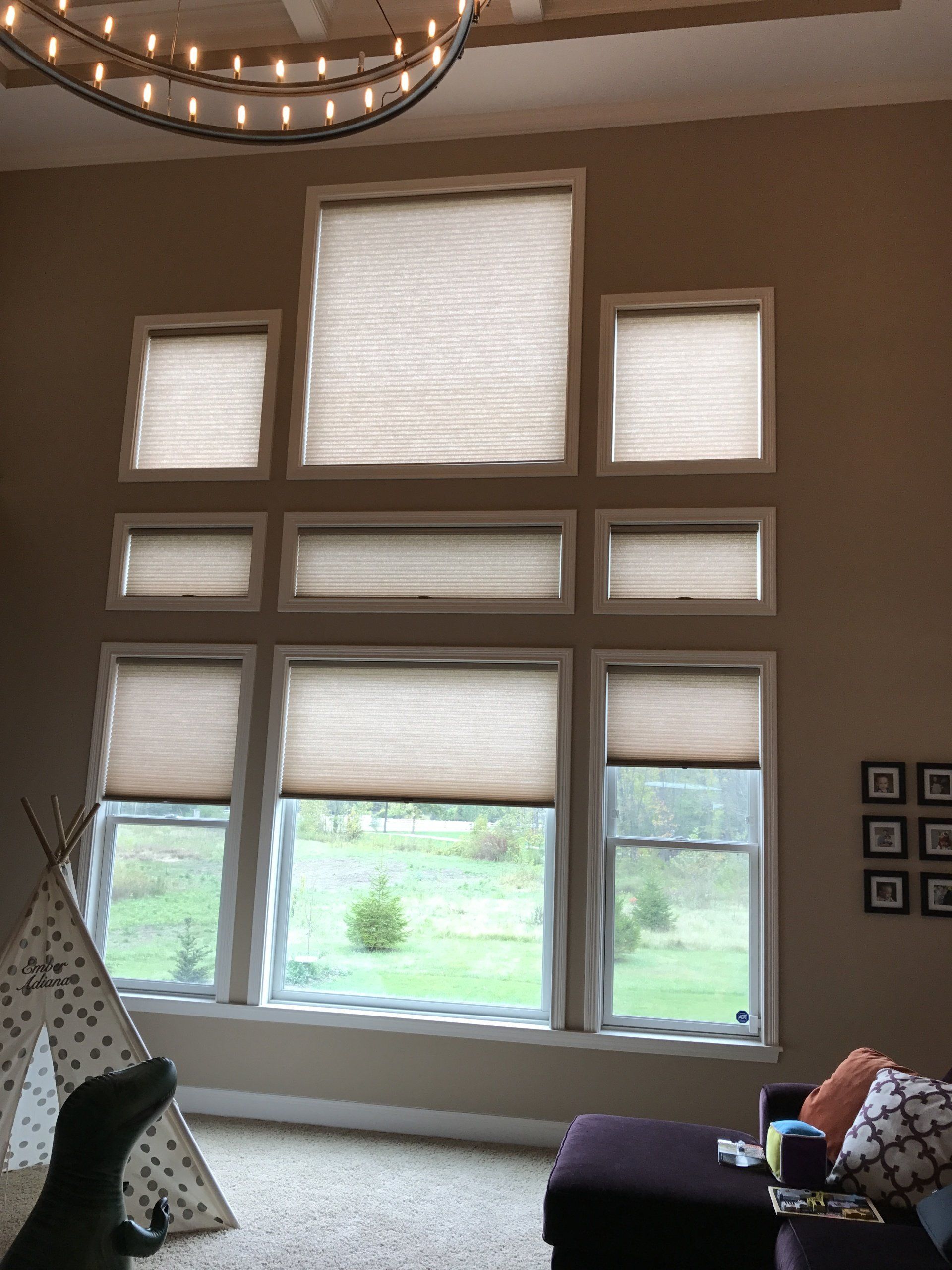 A wall with light brown paint features a large, multi-pane window configuration, all fitted with light-colored, pleated motorized shades. The bottom three windows are partially uncovered, revealing a green outdoor landscape with grass and trees. Above them, three rectangular windows are fully covered by the shades. The topmost large square window is also fully covered. A large, circular chandelier with multiple lights hangs from the ceiling. In the bottom left, a teepee and a dark green dinosaur toy are visible. On the right, a purple ottoman and a section of a couch with throw pillows are present.