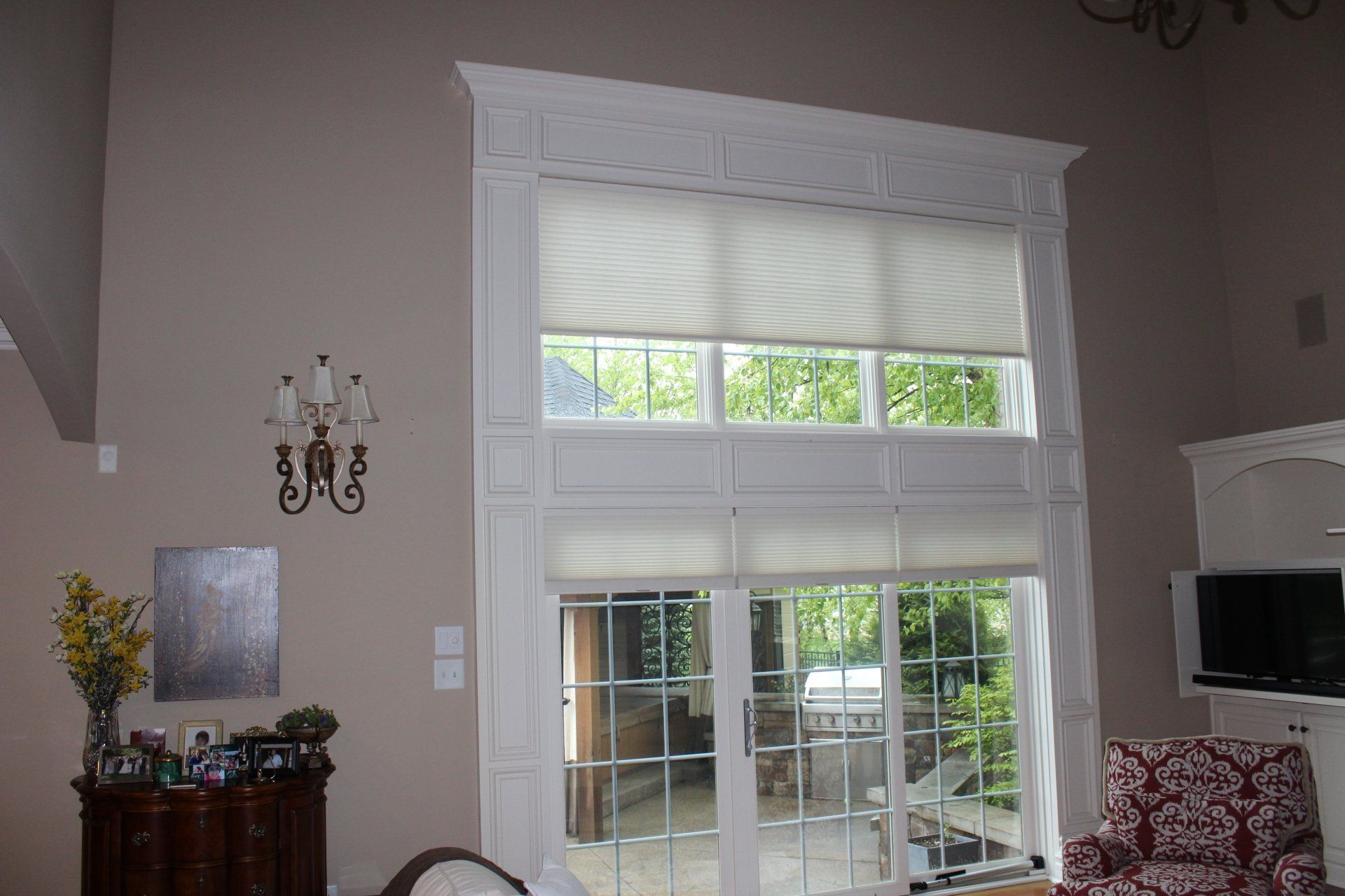 A large, multi-story window with intricate white trim is fitted with light-colored, pleated motorized shades. The top section and middle section of the window are covered by these shades, while the lower section, consisting of French doors with glass panes, is uncovered, revealing a patio area and green trees outside. A dark wooden chest of drawers with yellow flowers and framed photos sits to the left of the window, beneath a decorative wall sconce. On the right, a television is integrated into a white built-in unit, next to a chair with a red and white pattern. The walls are a light beige color.
