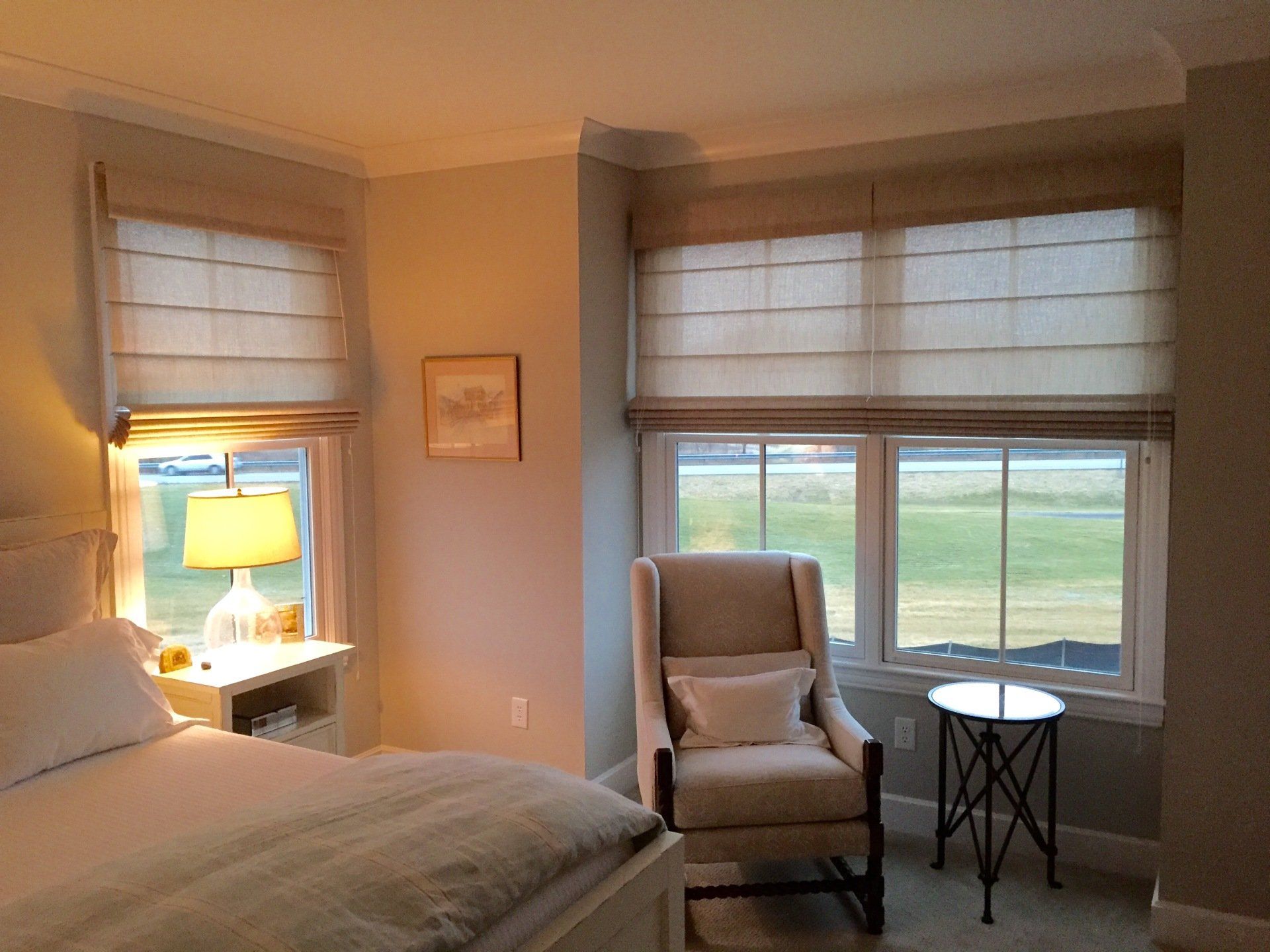 A cozy bedroom is illuminated by soft light, featuring two windows with tan roman shades drawn across them. On the left, a bedside table with a lamp casts a warm glow, next to a bed with white linens. On the right, a comfortable-looking armchair with a small pillow is positioned beside a round, dark-colored side table. 
