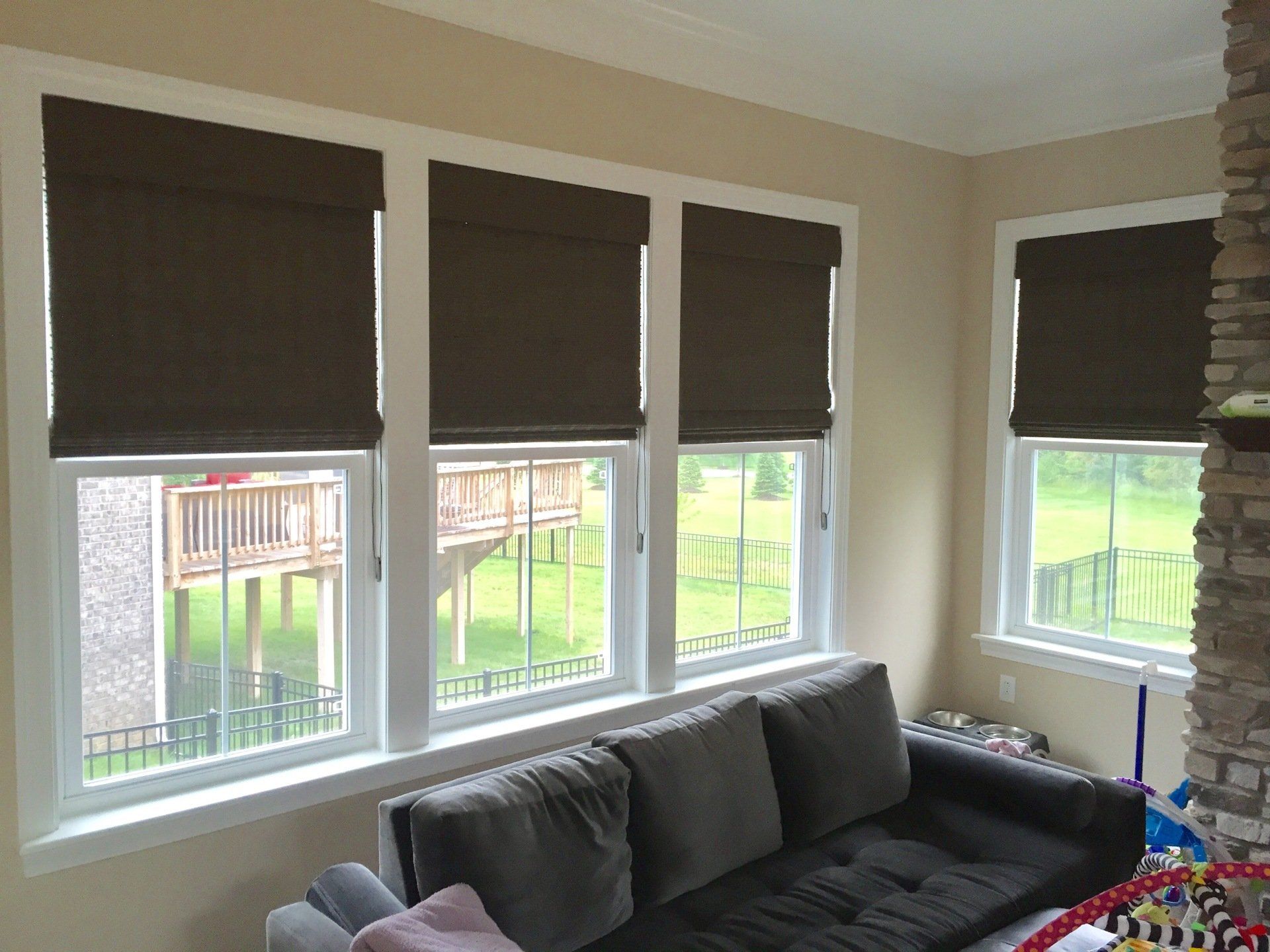 Four large windows are visible in a room with light beige walls. The top portion of each window is covered by dark brown room darkening shades, which appear to be Roman shades. The lower sections of the windows are uncovered, revealing a green outdoor landscape with a wooden deck structure, a metal fence, and grass. A dark gray sofa with throw pillows is positioned in front of the three leftmost windows. To the far right, a stone-faced column or fireplace is partially visible, with colorful items suggesting children's toys near its base.