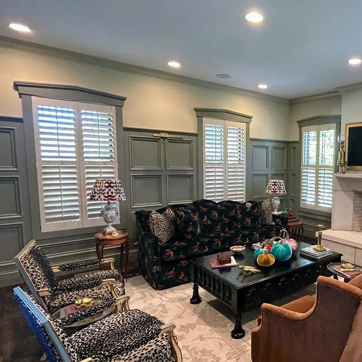 A well-appointed living room featuring white plantation shutters on three windows, framing a seating area with a patterned sofa, armchairs, and a coffee table.